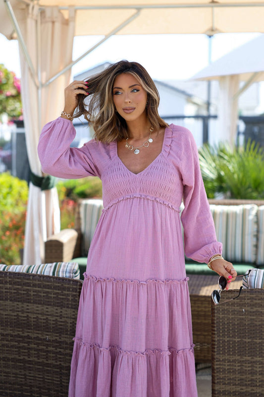 Woman wearing the Rosy Pink Smock Tiered Midi Dress stands outdoors, touching her hair with patio furniture and greenery behind her.