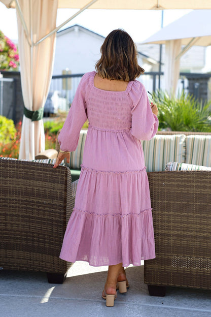A woman in a Rosy Pink Smock Tiered Midi Dress stands with her back to the camera by outdoor wicker furniture.