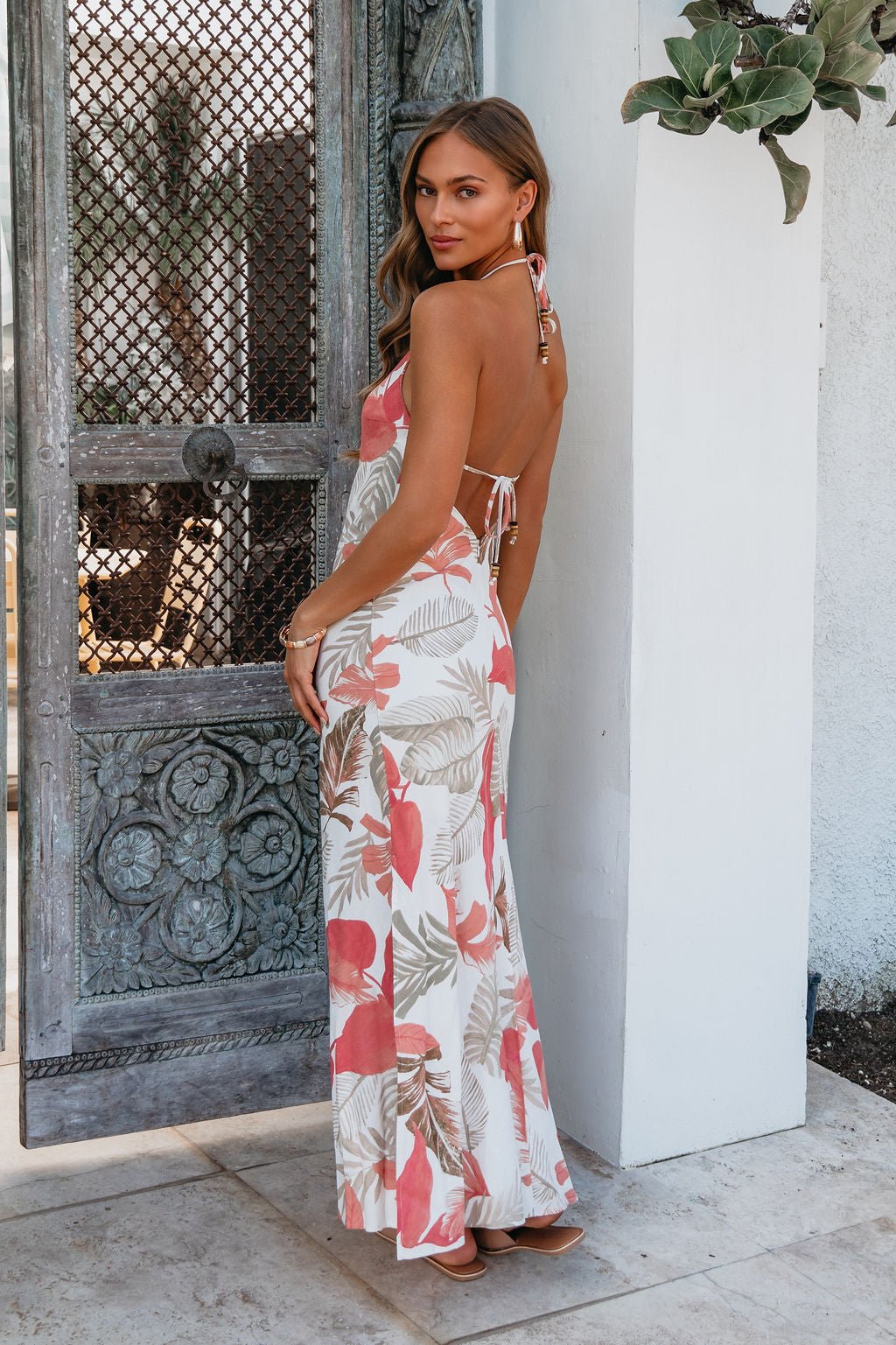 Woman in the Rosy Reef Escape Halter Maxi Dress stands by an ornate wooden door, exuding effortless resort style.