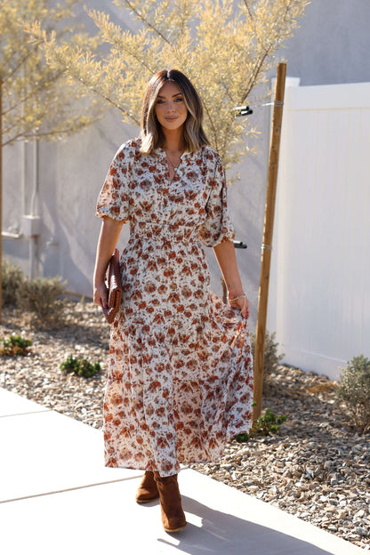 Woman wearing the Rowan Cream & Rust Tiered Midi Dress, walking outside among plants and a fence on a sunny day.