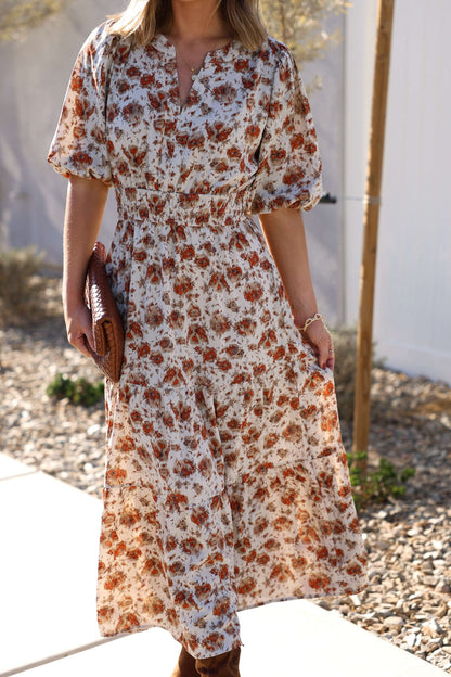 A woman in the Rowan Cream & Rust Tiered Midi Dress holds a woven clutch while standing outdoors on a sunny day.