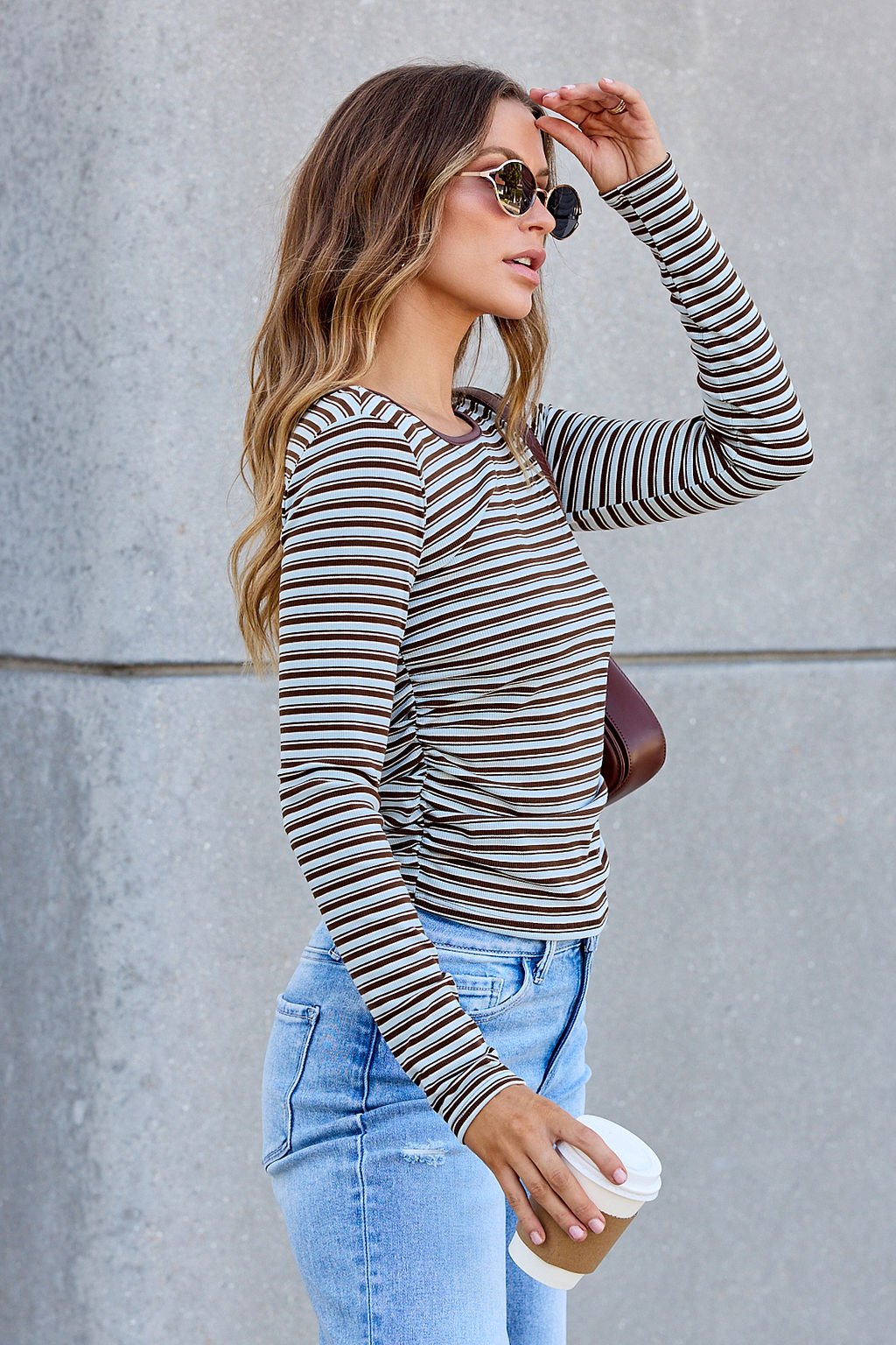Woman in sunglasses wears the Ruched Stripe Top - Brown with jeans, holding coffee against a concrete wall.