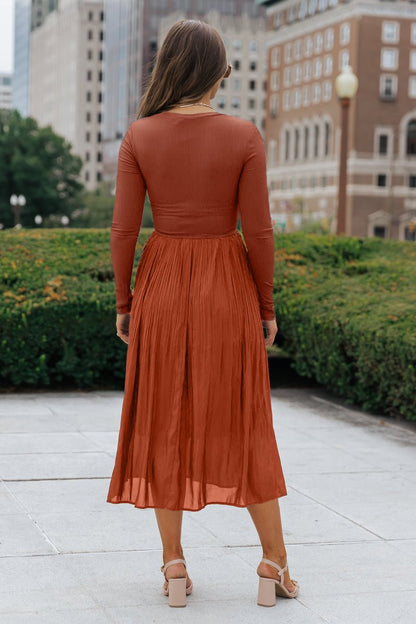 Woman in a Rust Contrast Pleated Midi Dress with ribbed details stands outside, pleated skirt flowing as she faces buildings and greenery.