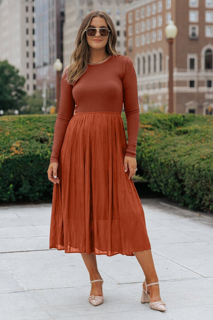 Woman in sunglasses wears a Rust Contrast Pleated Midi Dress and nude heels, walking on a city sidewalk with greenery behind her.