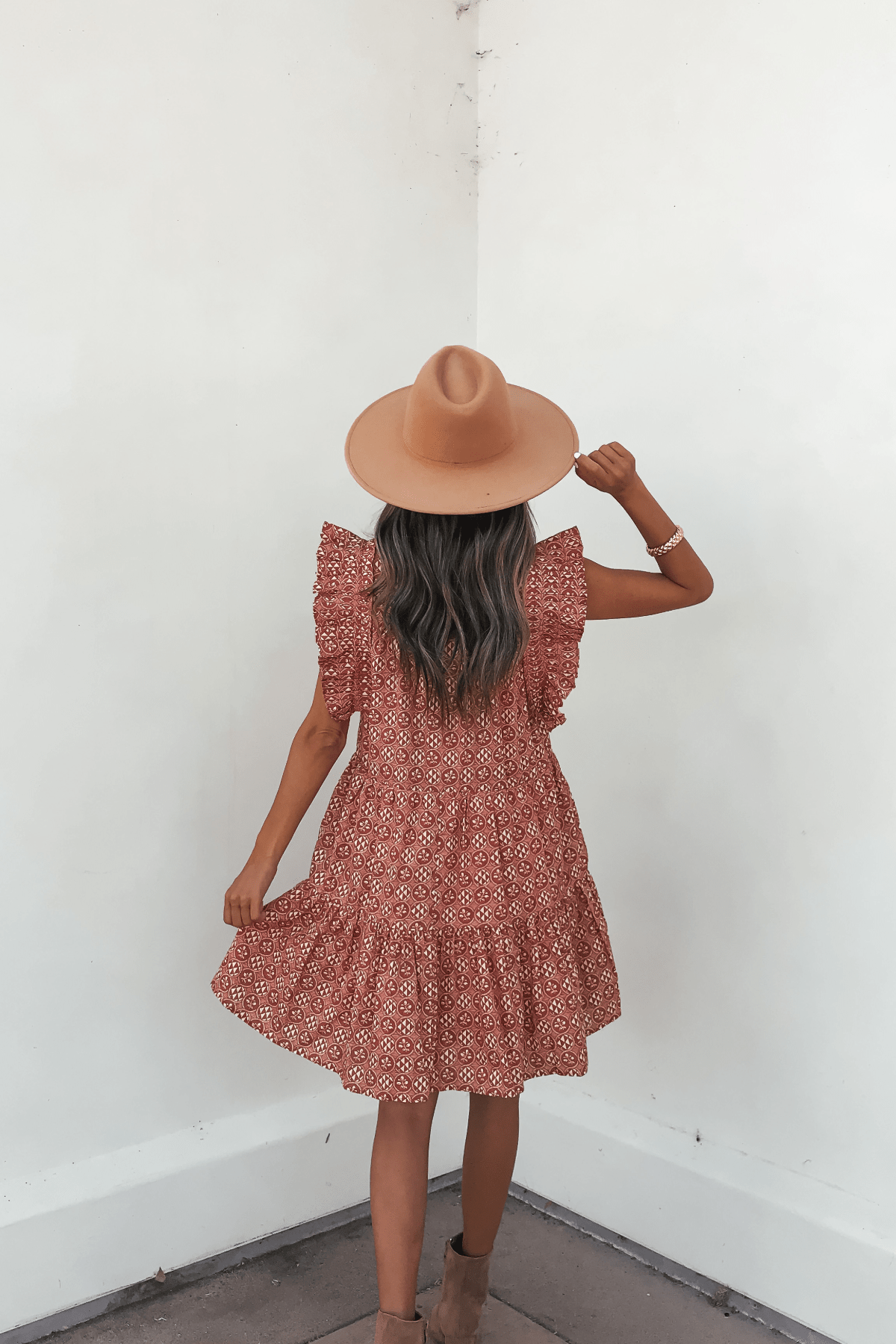 A woman in the Rust Print Flutter Sleeve Mini Dress stands by a white wall, holding her hat and wearing tan ankle boots.