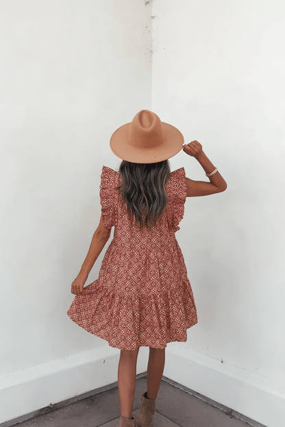 A woman in the Rust Print Flutter Sleeve Mini Dress stands by a white wall, holding her hat and wearing tan ankle boots.