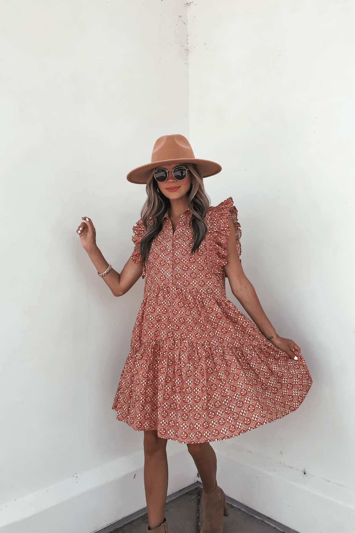 Woman in a Rust Print Flutter Sleeve Mini Dress - FINAL SALE, tan hat, and sunglasses stands by a white wall holding her dress.