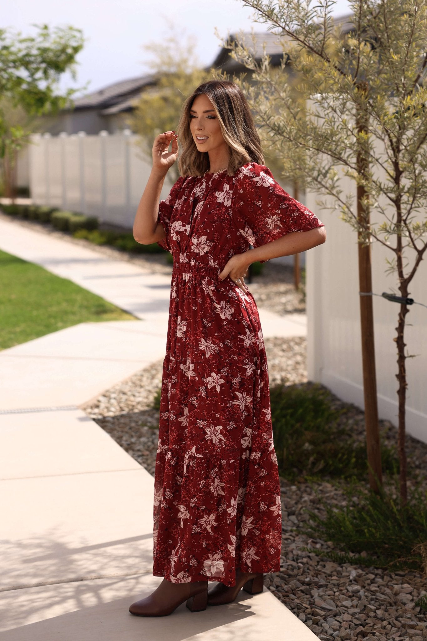 A woman wearing the Rustling Leaves Wine Floral Print Maxi Dress smiles on a sunny suburban sidewalk with grass and trees.