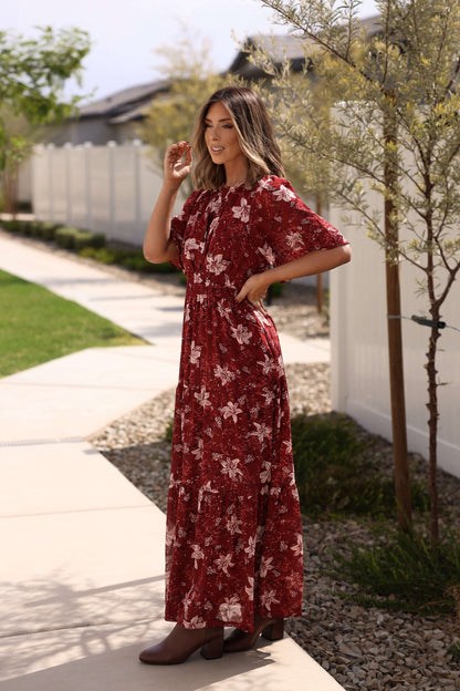 A woman wearing the Rustling Leaves Wine Floral Print Maxi Dress smiles on a sunny suburban sidewalk with grass and trees.