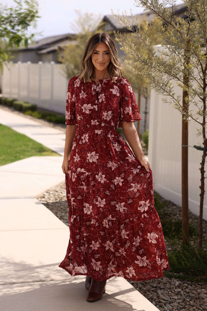 Woman smiles outdoors in the Rustling Leaves Wine Floral Print Maxi Dress, with greenery and a white fence nearby.