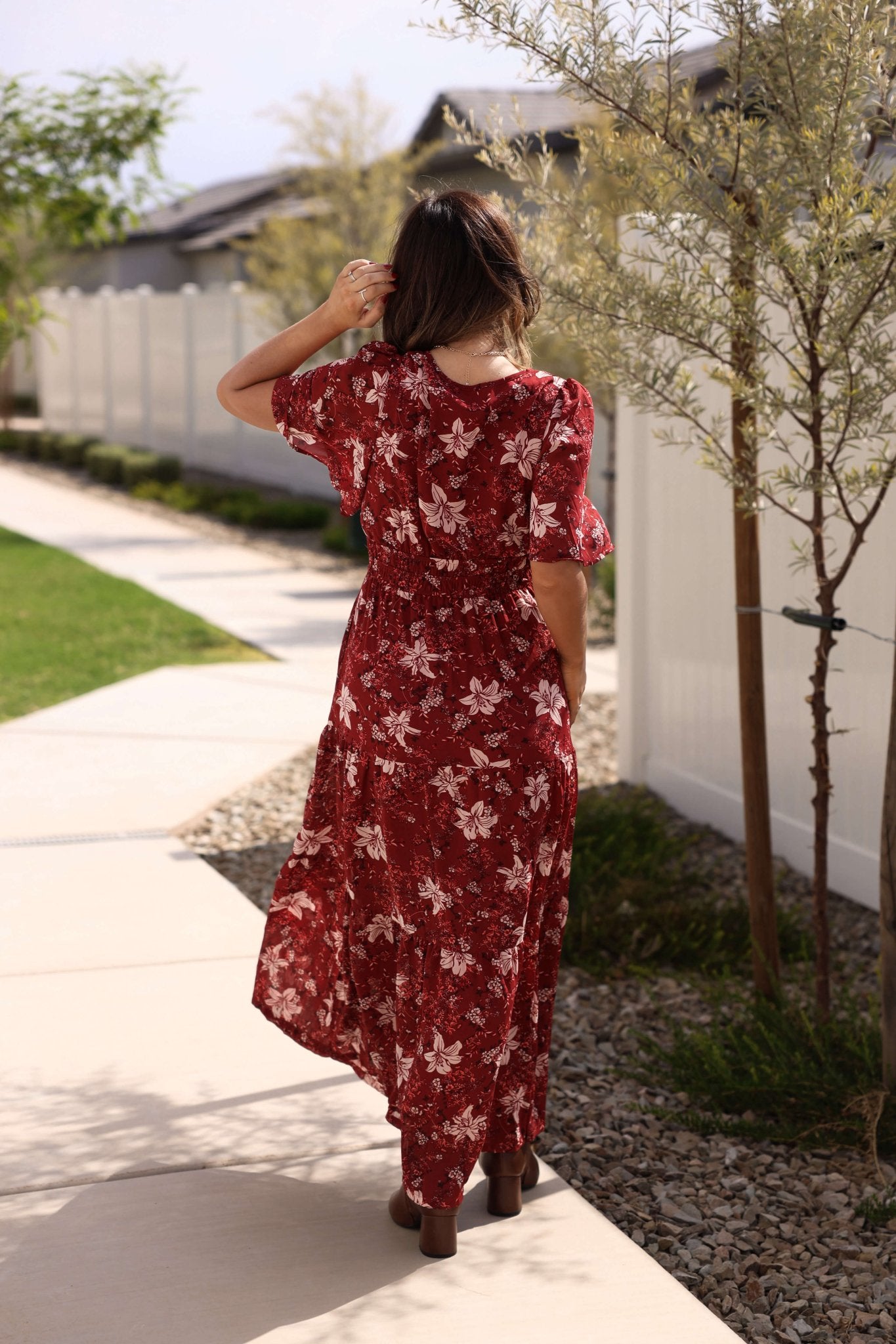 A woman in the Rustling Leaves Wine Floral Print Maxi Dress walks on a sidewalk, facing away, hand in her hair.