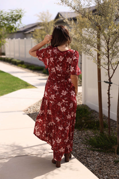 A woman in the Rustling Leaves Wine Floral Print Maxi Dress walks on a sidewalk, facing away, hand in her hair.
