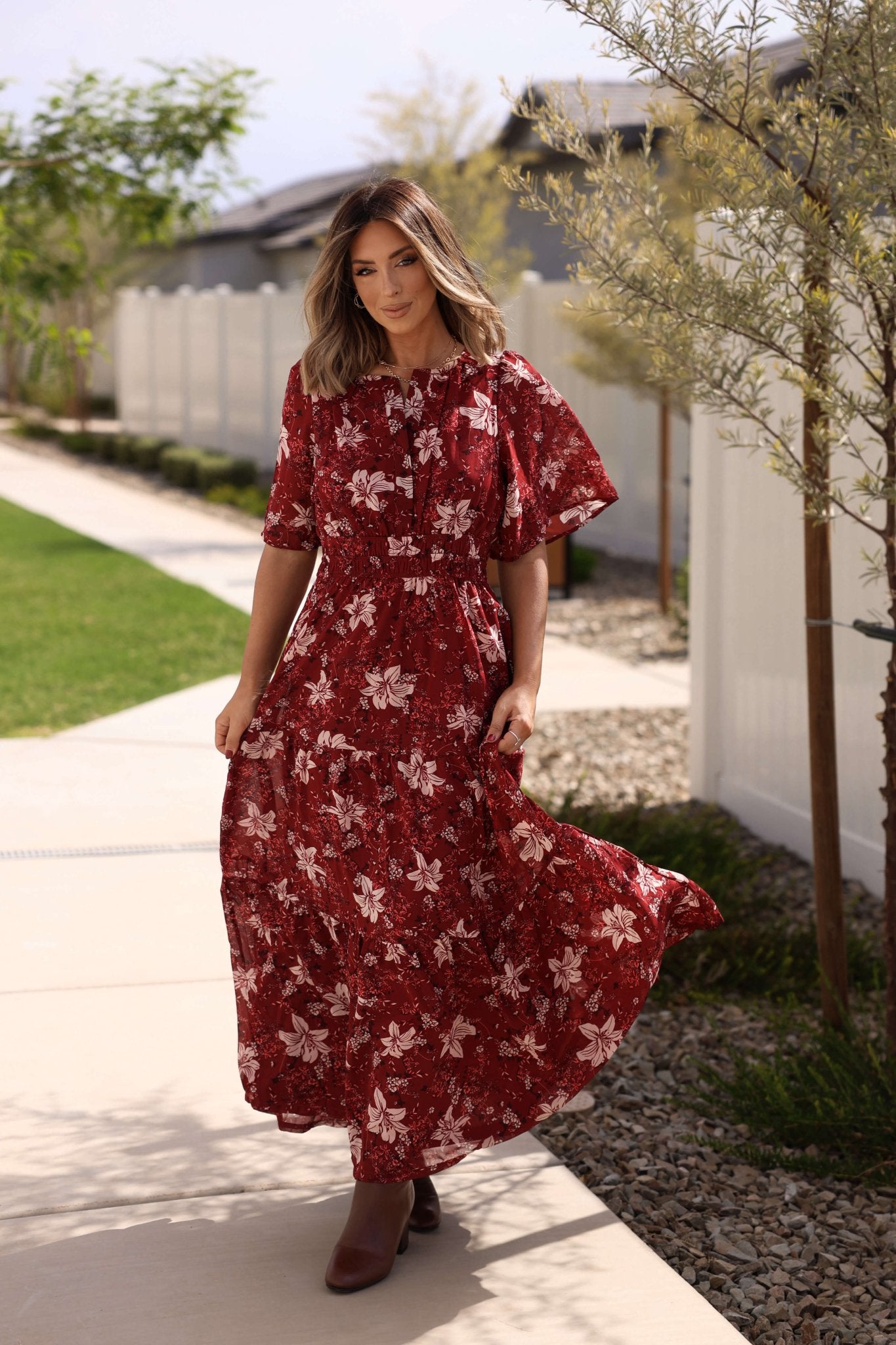 A woman in the Rustling Leaves Wine Floral Print Maxi Dress strolls on a sunny sidewalk near a white fence and green lawn.
