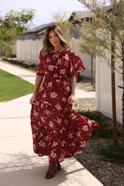 A woman in the Rustling Leaves Wine Floral Print Maxi Dress strolls on a sunny sidewalk near a white fence and green lawn.