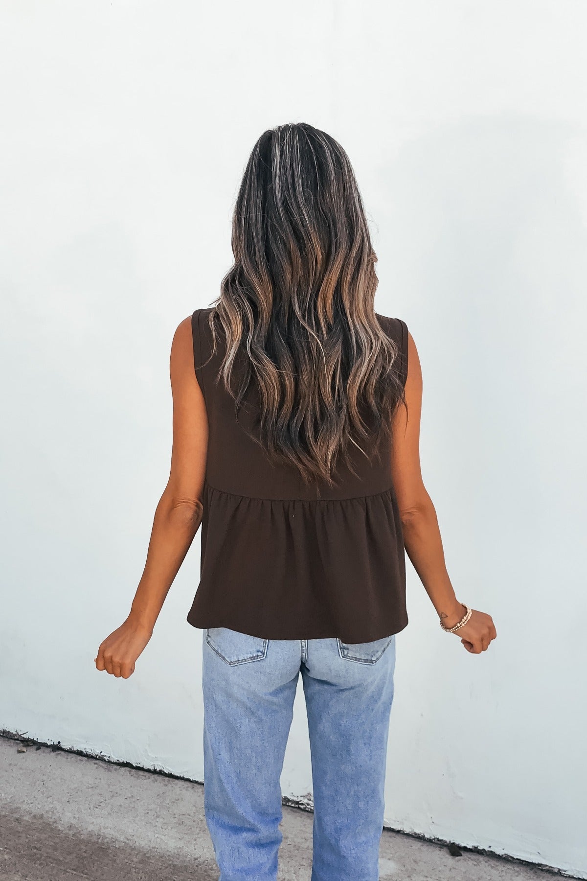 Woman with long wavy hair in a relaxed fit Sable Pleated Top - Brown and light blue jeans, facing a white wall.