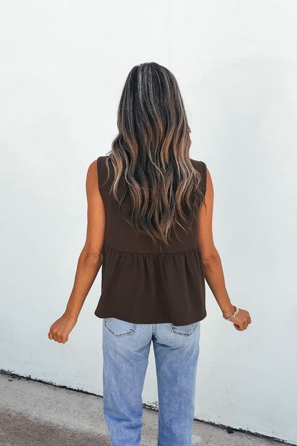 Woman with long wavy hair in a relaxed fit Sable Pleated Top - Brown and light blue jeans, facing a white wall.