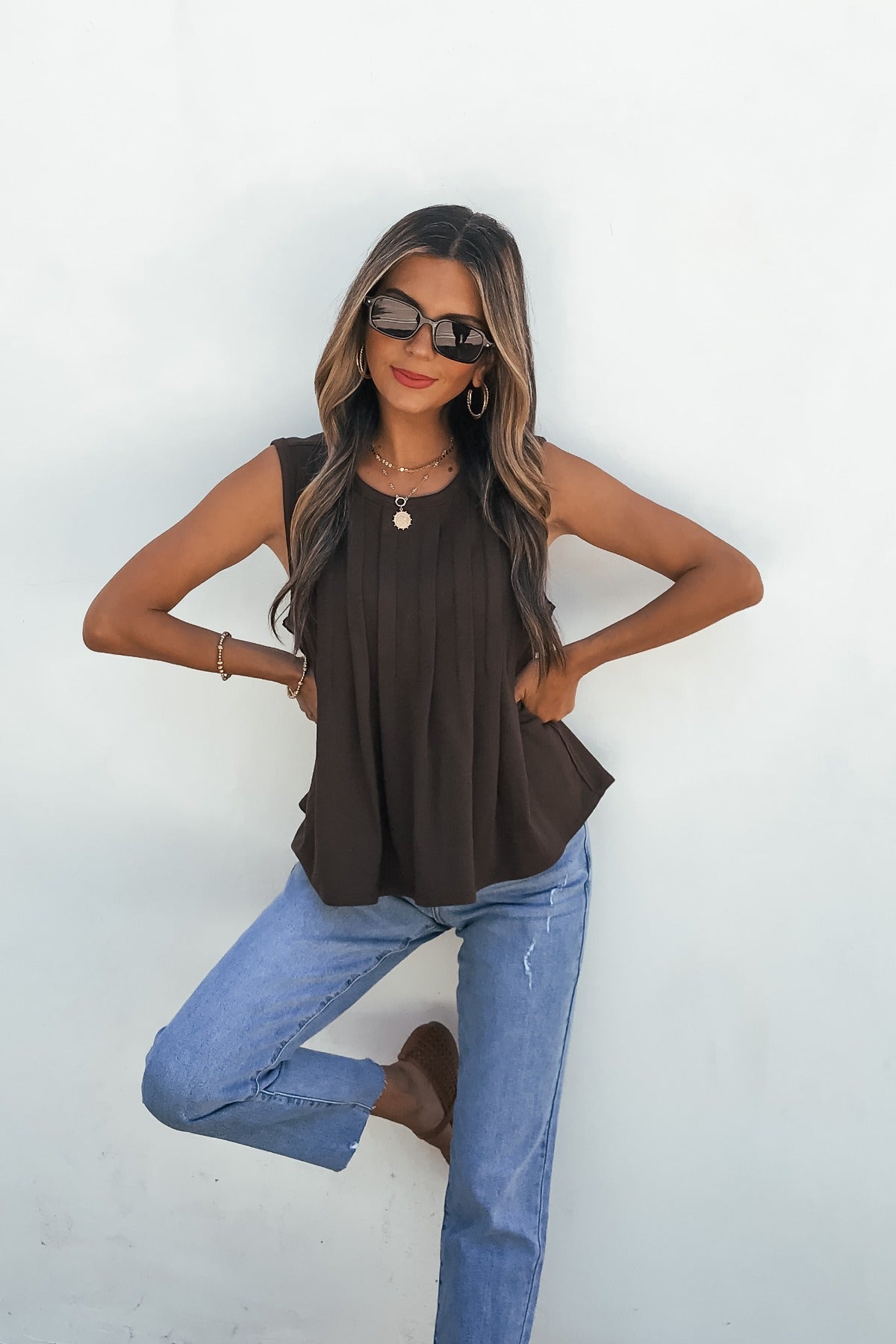 Woman poses in sunglasses, Sable Pleated Top - Brown, relaxed blue jeans, brown shoes, one knee raised by a white wall.