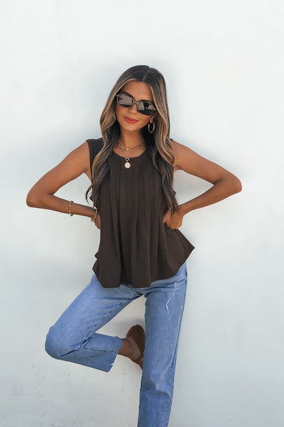 Woman poses in sunglasses, Sable Pleated Top - Brown, relaxed blue jeans, brown shoes, one knee raised by a white wall.