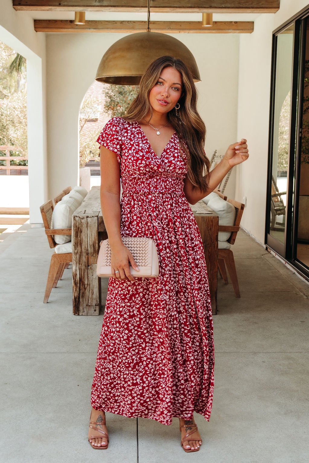 Woman wears Sabrina Red and White Floral Maxi Dress - DOORBUSTER, holding a beige clutch in a chic outdoor dining space.