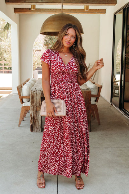 Woman wears Sabrina Red and White Floral Maxi Dress - DOORBUSTER, holding a beige clutch in a chic outdoor dining space.