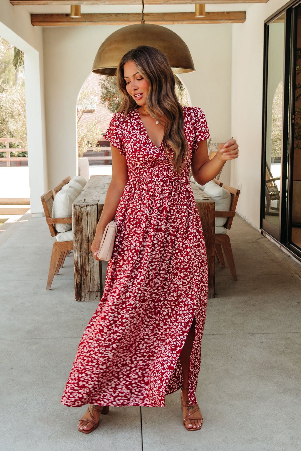 Woman in the Sabrina Red and White Floral Maxi Dress - DOORBUSTER holds a beige clutch on a patio with wooden furniture and lights.