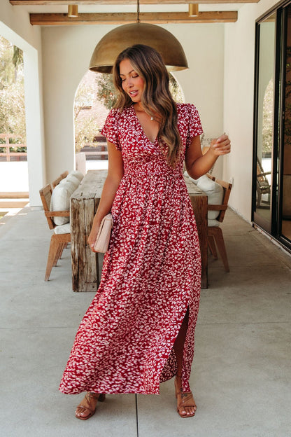 Woman in the Sabrina Red and White Floral Maxi Dress - DOORBUSTER holds a beige clutch on a patio with wooden furniture and lights.