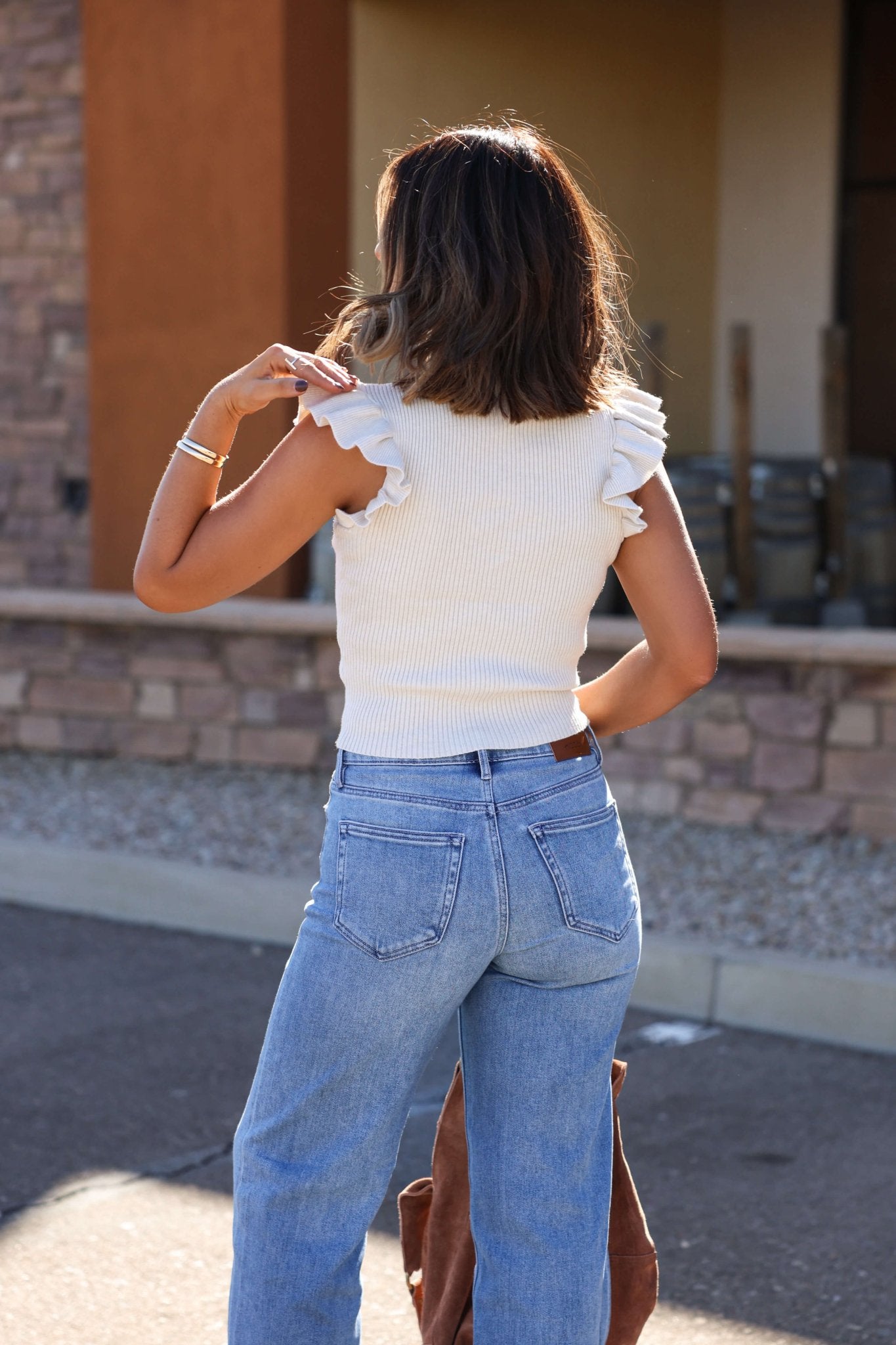 A woman stands outdoors with her back to the camera, wearing the Sadie and Sage Cream Ruffle Sweater and blue jeans.