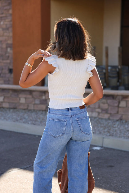 A woman stands outdoors with her back to the camera, wearing the Sadie and Sage Cream Ruffle Sweater and blue jeans.