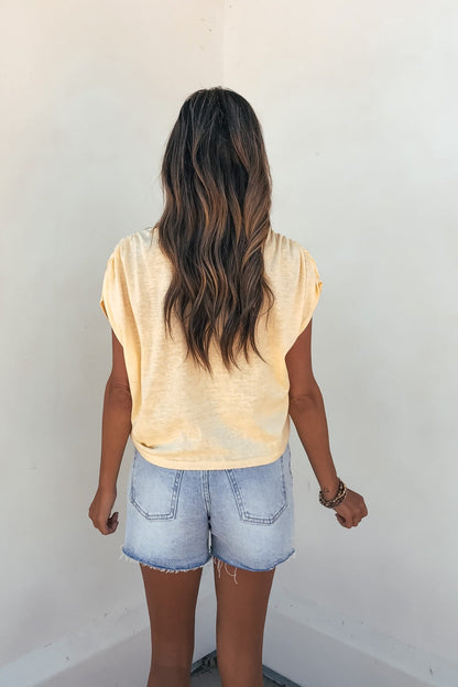 A woman with long wavy hair faces a plain wall, wearing a yellow Sadie and Sage Marlow Gathered Top and light denim shorts.