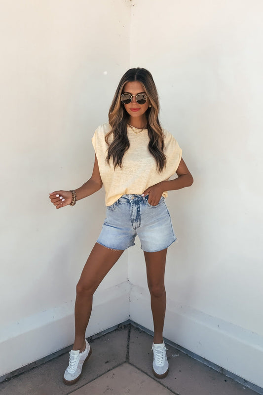 Woman in sunglasses, denim shorts, and white sneakers wears the Sadie and Sage Marlow Gathered Top - Yellow against an off-white wall.