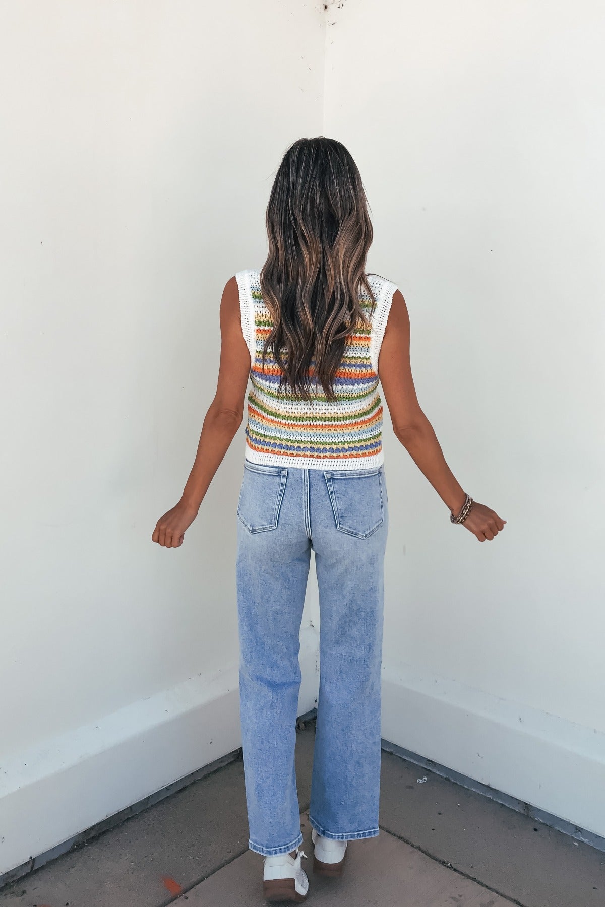 A woman in a Sadie and Sage Stripe Knit Sweater Tank Top and light blue jeans faces a white wall, back to the camera.