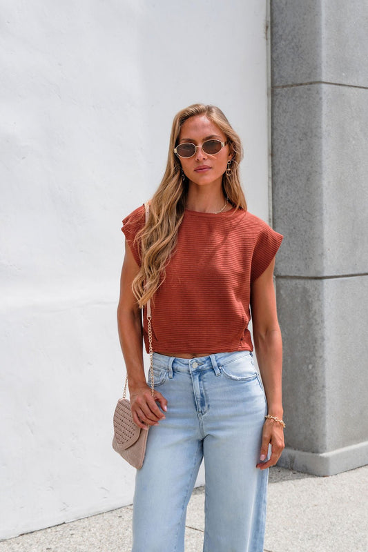 Woman in sunglasses wears the Sadie & Sage Ribbed Top - Rust, light jeans, and a textured bag by a white wall and gray pillar.