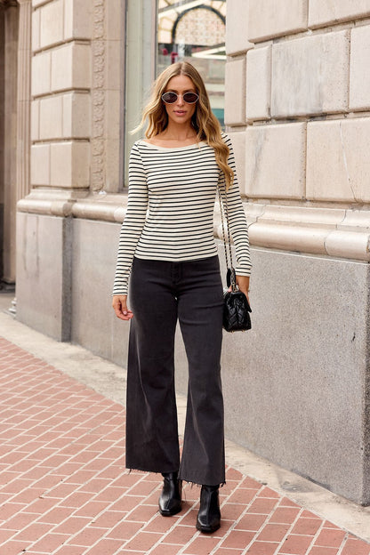 Woman in sunglasses wears the Sadie & Sage Striped Boatneck Top - Cream, black wide-leg pants, and ankle boots while strolling downtown.