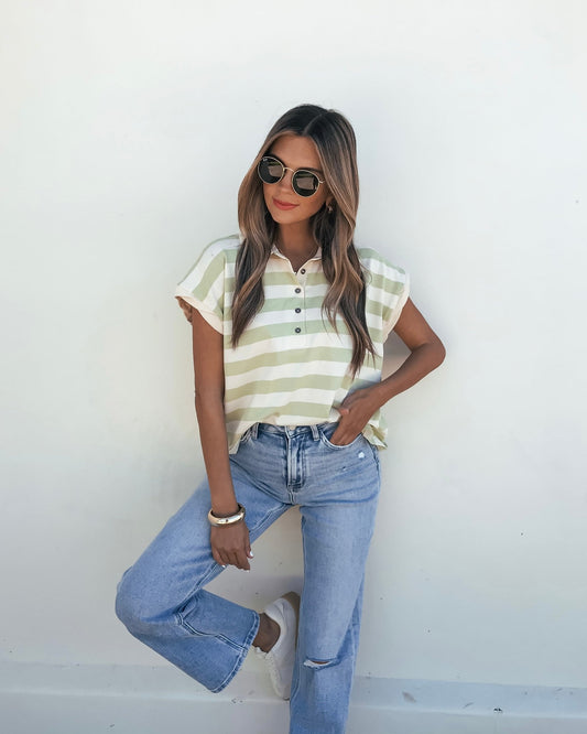 Woman in sunglasses models the Sage and Ivory Striped Button Up Shirt with blue jeans, posing casually against a white wall.