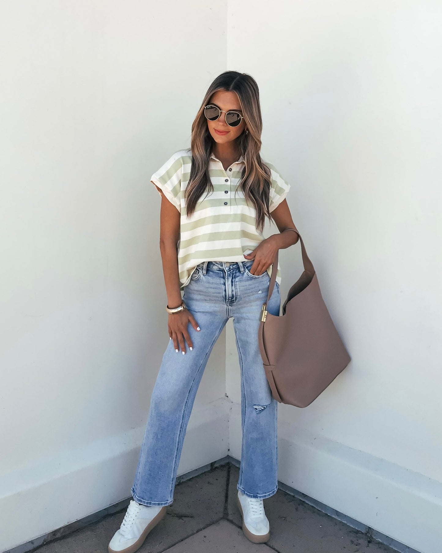 Woman in sunglasses wearing a Sage and Ivory Striped Button Up Shirt, light blue jeans, white sneakers, and a tote bag by a white wall.