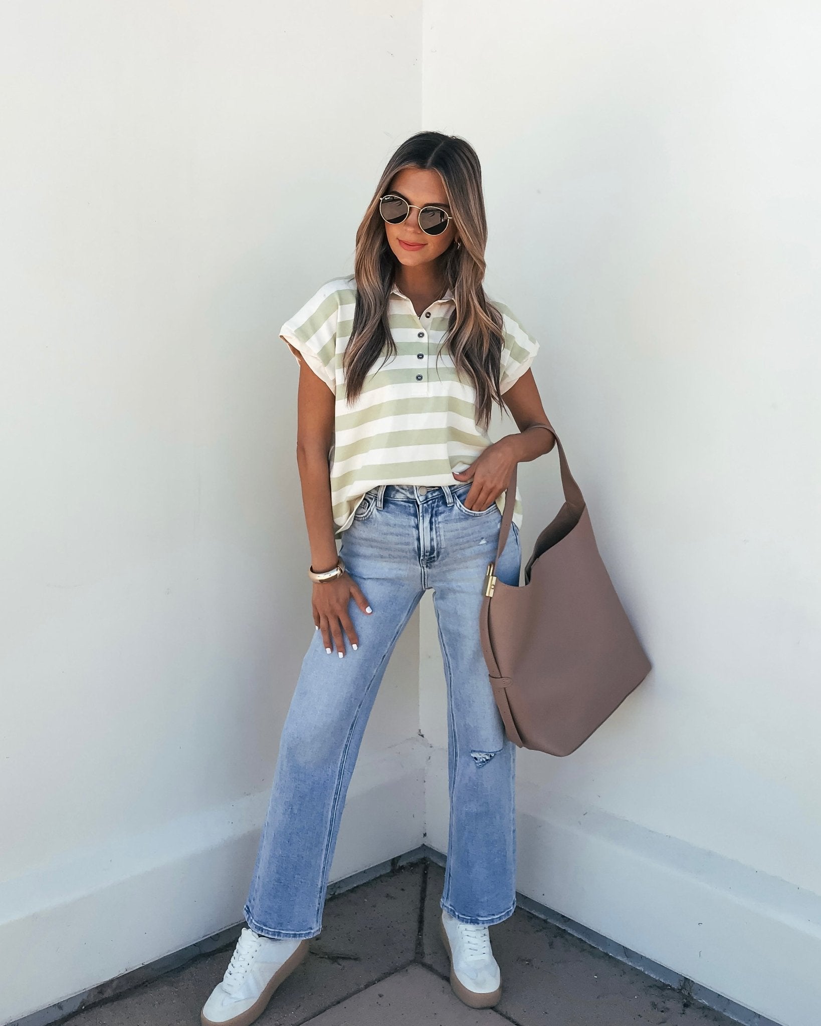 Woman in sunglasses wearing a Sage and Ivory Striped Button Up Shirt, light blue jeans, white sneakers, and a tote bag by a white wall.
