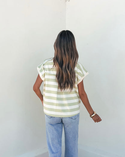 A woman in the Sage and Ivory Striped Button Up Shirt and jeans stands with her back to the camera, facing a white wall.