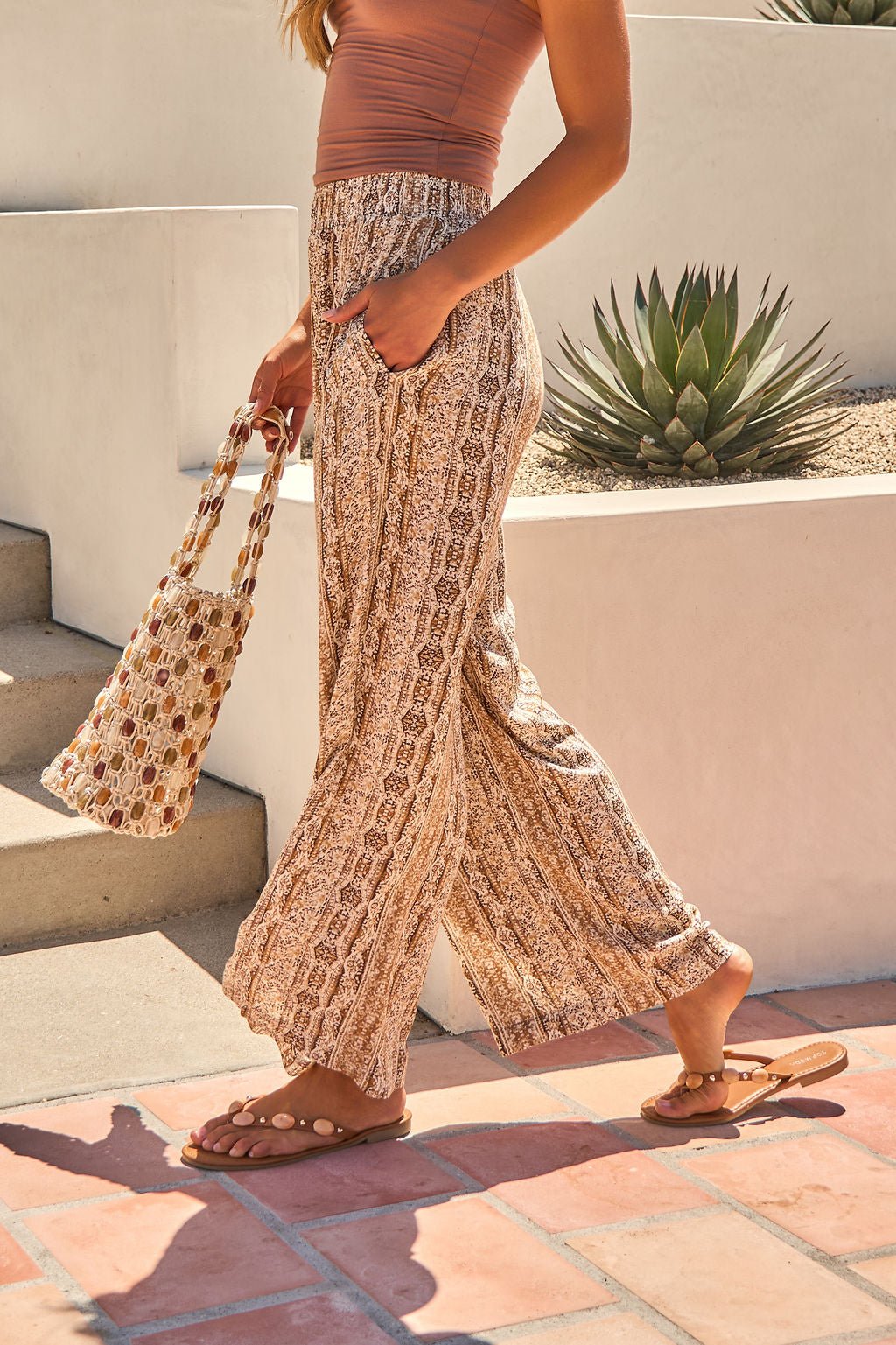 Woman outdoors in Sahara Smocked Palazzo Pants - Brown and tank top, holding a woven tote near desert plants and steps.