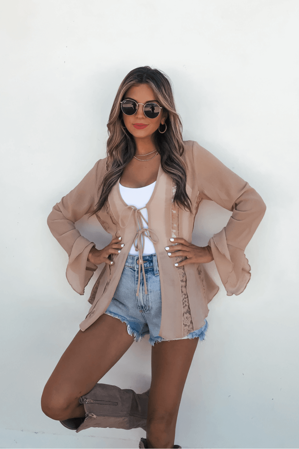 Woman in sunglasses, denim shorts, knee-high boots, and a Sand Bell Sleeve Sheer Lace Top poses against a white wall.