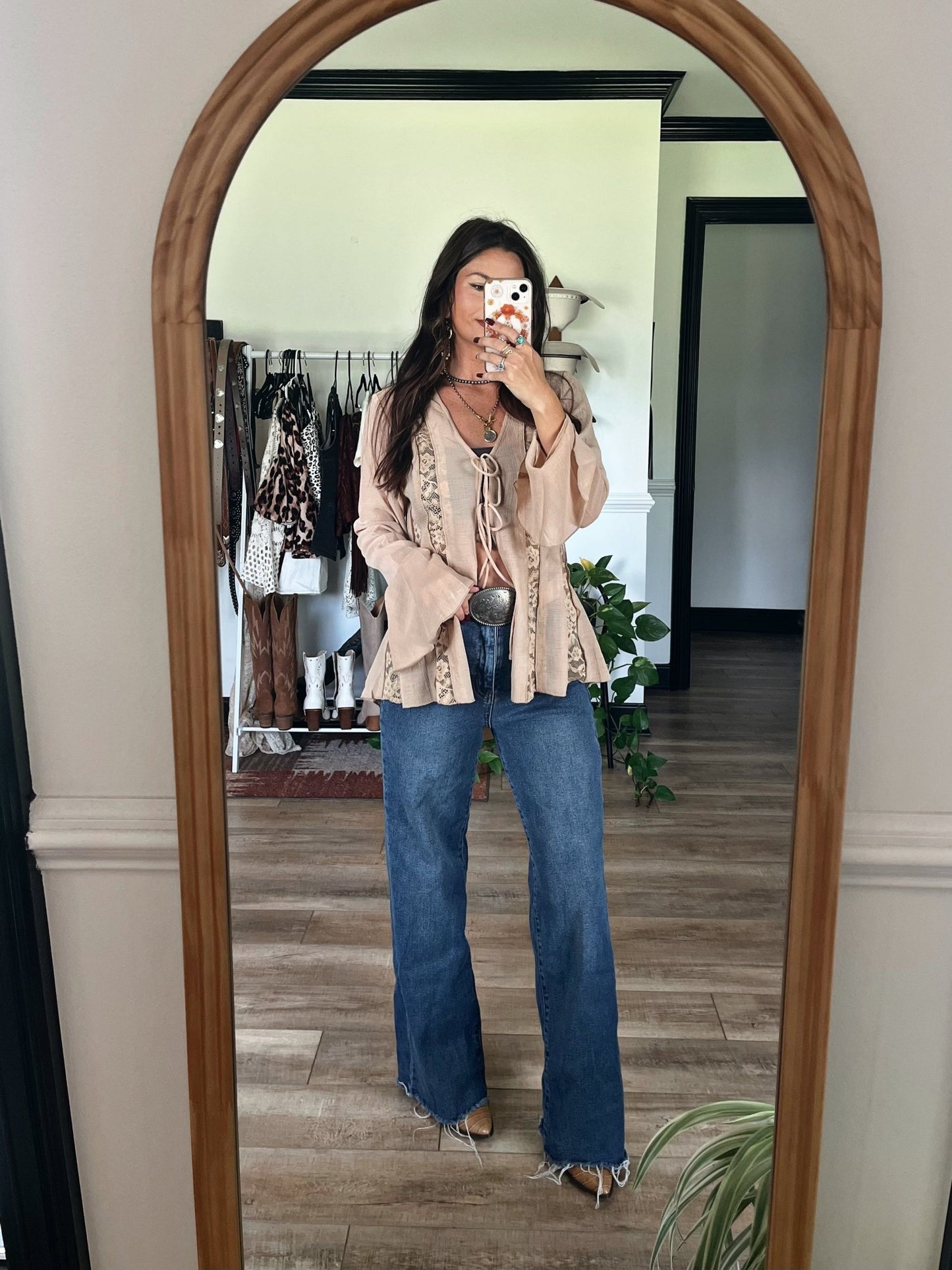 Woman taking a mirror selfie in a Sand Bell Sleeve Sheer Lace Top and wide-leg jeans, clothes on a rack in the background.