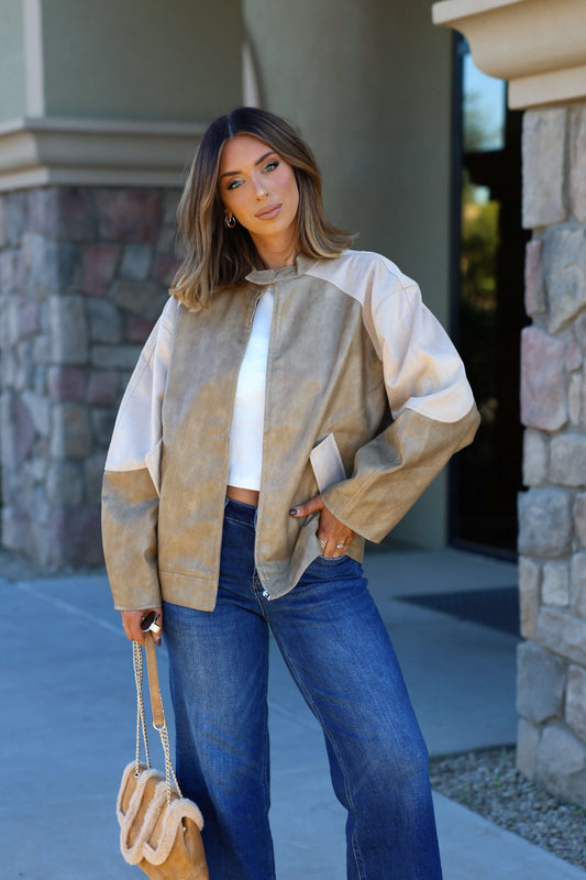 A woman in a Sand Faux Leather Loose Suede Jacket stands outside holding a tan handbag near a stone building.