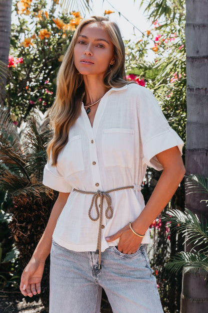 A woman in a Santorini Ivory Belted Linen Top and jeans stands outside among palm trees and vibrant flowers.