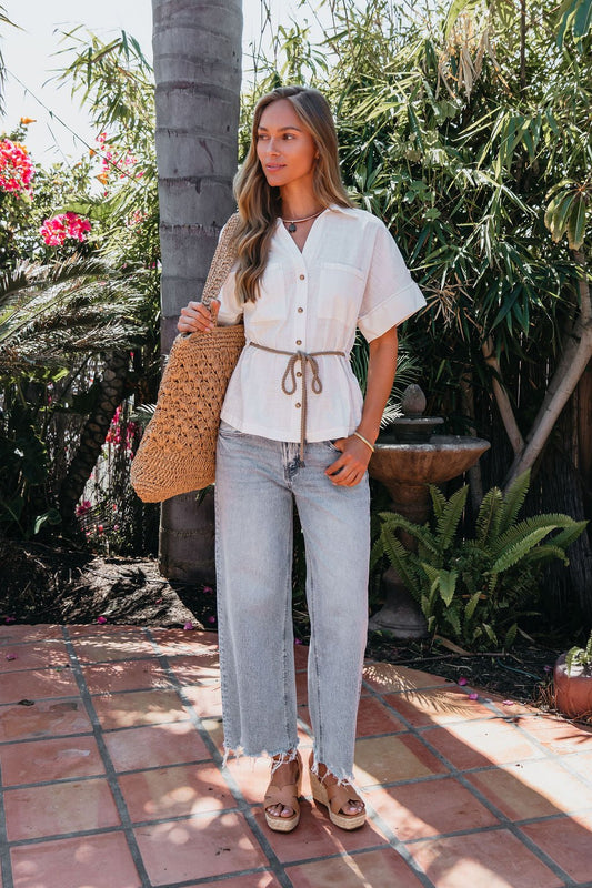 Woman outdoors in the Santorini Ivory Belted Linen Top, belted waist, light jeans, sandals, and a large woven bag.