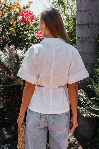 A woman in a Santorini Ivory Belted Linen Top and light jeans stands outdoors, surrounded by lush greenery and flowers.