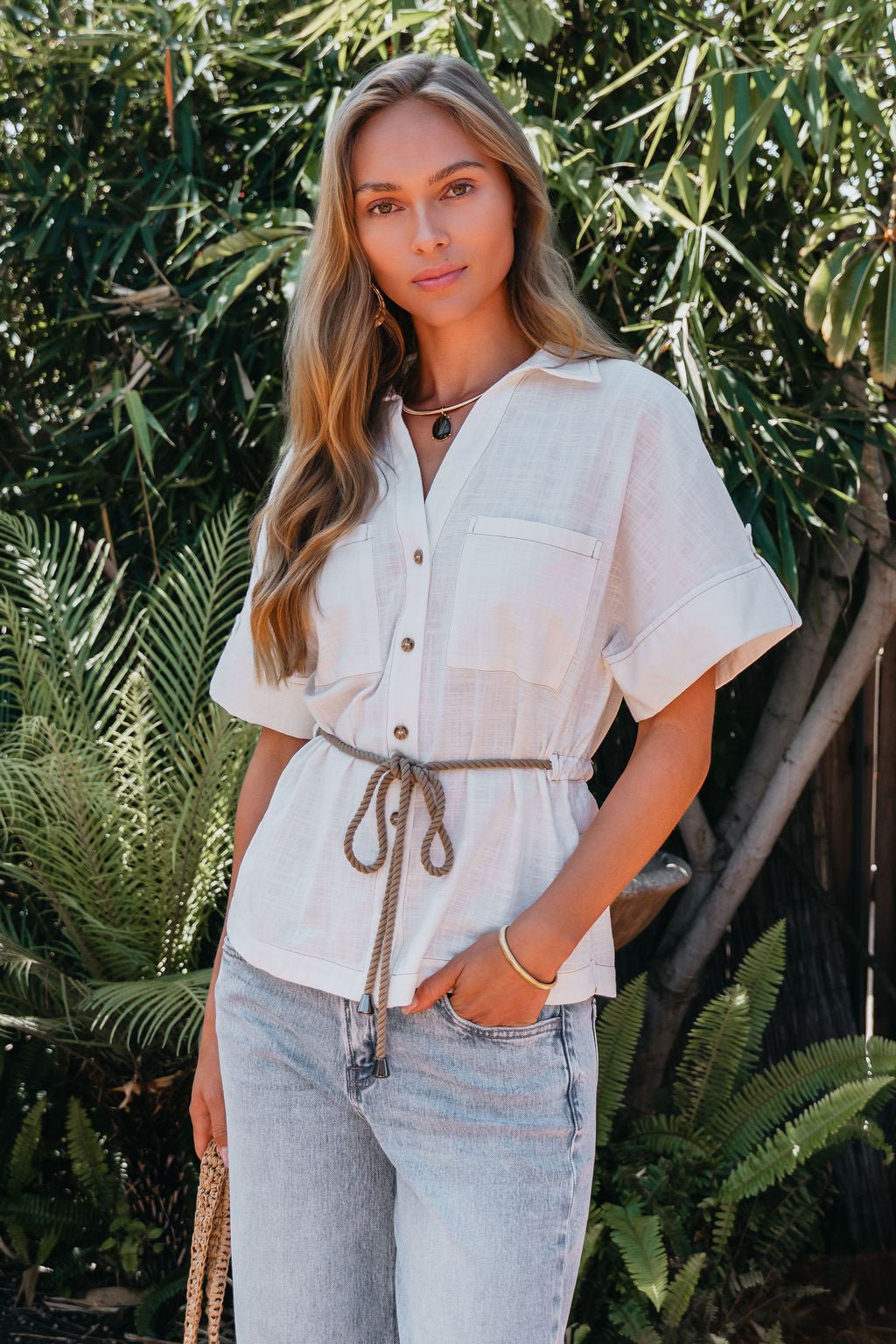 A woman stands outdoors by green plants, wearing a Santorini Ivory Belted Linen Top and jeans.