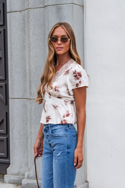 Woman in sunglasses wears a Satin Lace Floral Print Top - Cream and blue jeans by a gray stone building, holding something.