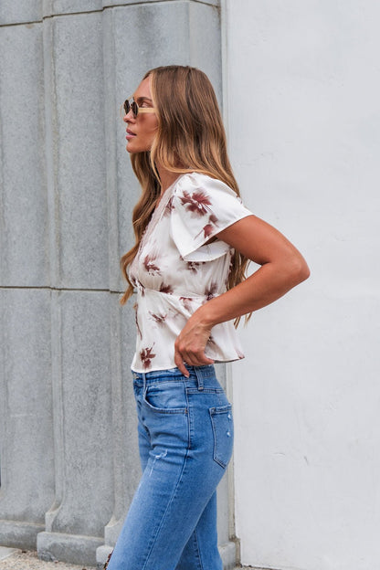 Woman in sunglasses, floral top, and jeans stands sideways by a stone wall, long hair flowing and hands on her hips.