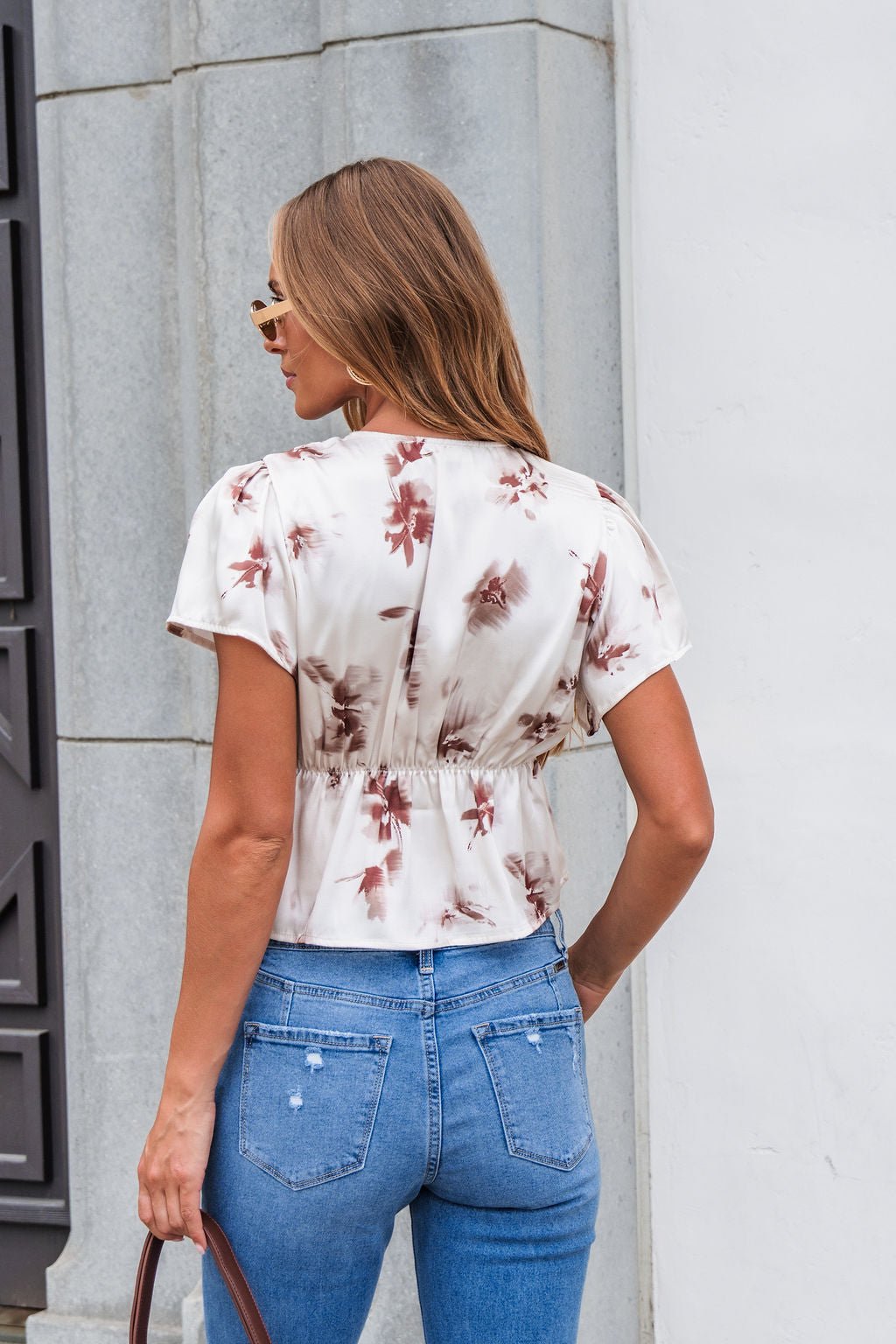 Woman in sunglasses and blue jeans wears a Cream Satin Lace Floral Print Top, standing outdoors with her back to the camera.