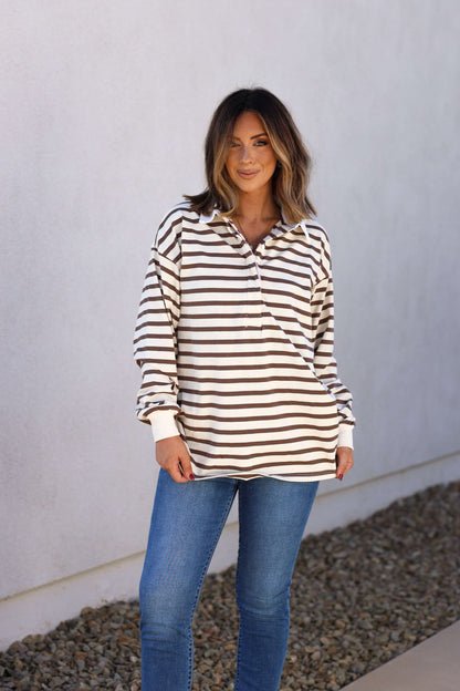 Woman in the Sawyer Brown Striped Pullover - DOORBUSTER and blue jeans stands on a stone path by a white wall, perfect for fall.