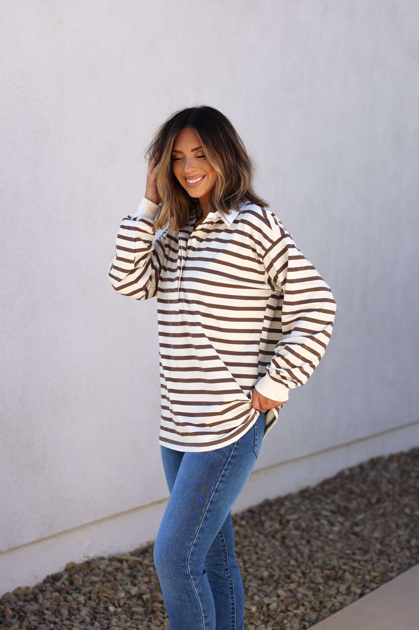 Woman smiles in the Sawyer Brown Striped Pullover - DOORBUSTER with blue jeans, styled effortlessly by a white wall and rocks.